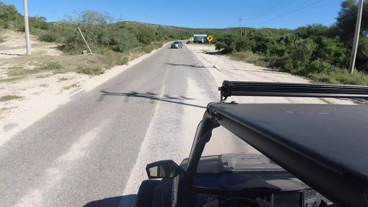 Los Cabos road scene one second in, with the black vehicle roof at lower right, a straight stretch of pavement, roadside scrub, utility poles, distant signs, and no visible people.