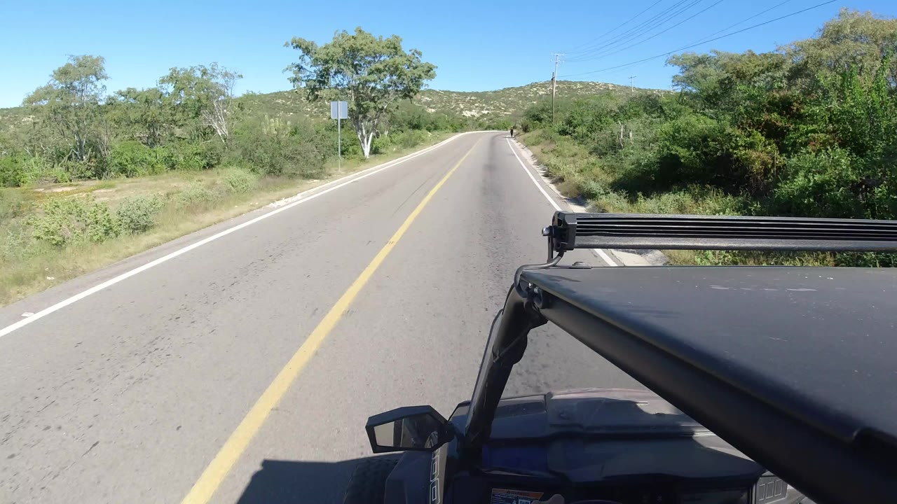 Opening frame of a Jeep-mounted road view in Los Cabos, Mexico, with a straight paved lane, green brush, low hills, clear sky, and no visible people.
