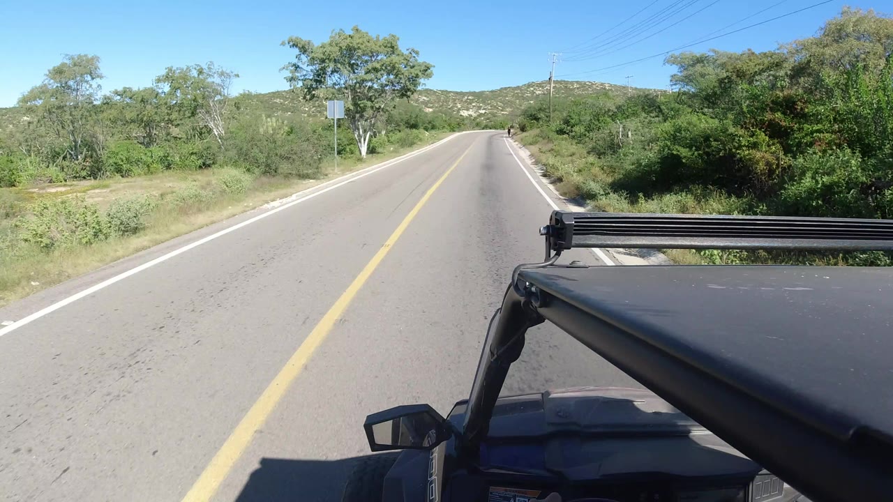 Vehicle-mounted view in Los Cabos, Mexico, showing the Jeep moving along a straight two-lane road bordered by dense green vegetation and no visible people.
