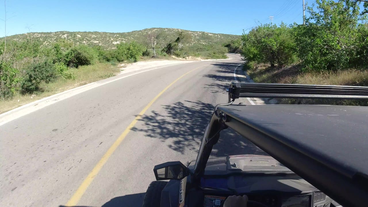 The vehicle-facing view continues along the Los Cabos road, with the hood and roof bar at lower right, scrub vegetation on both sides, distant hills, and no visible people.