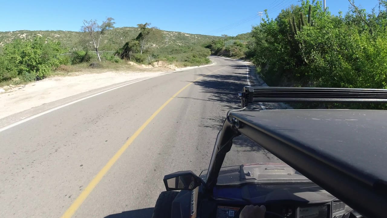 Road view curving ahead past green brush and a sandy shoulder.