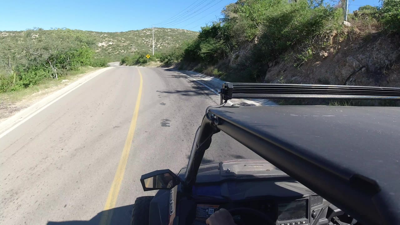 Los Cabos road POV with the black vehicle roof and light bar in the foreground, pale pavement ahead, dense green brush to the right, and no visible people.