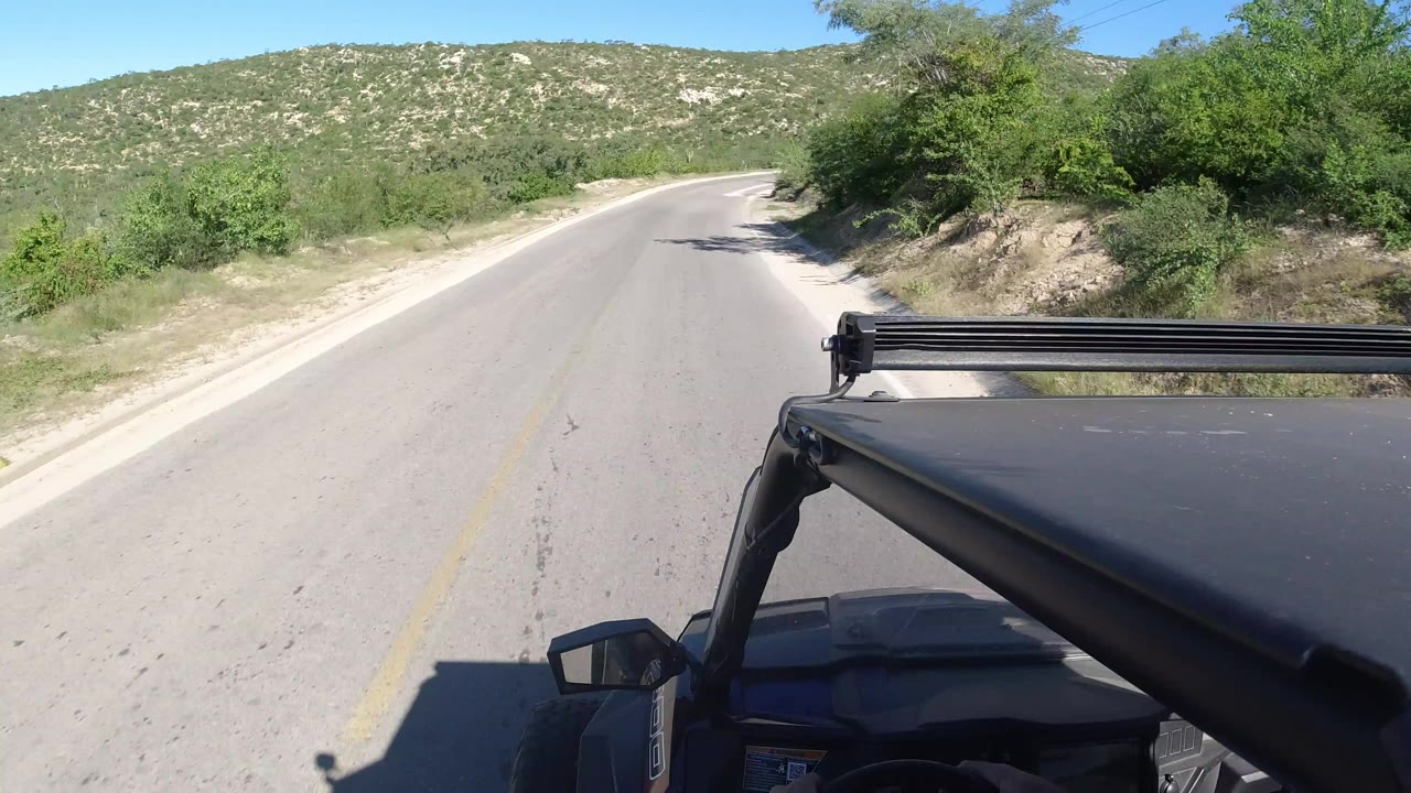 A Los Cabos road curves ahead through scrubby green hills, with the black vehicle hood and roof rack visible in the foreground and no visible people.