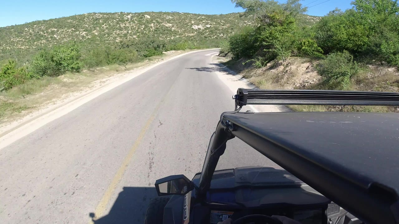 The vehicle continues along a quiet Los Cabos road, with the hood and mirror at bottom frame, pale pavement ahead, green brush, and no visible people.