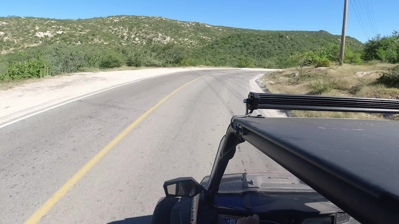 A Los Cabos rural road continues ahead from a vehicle-mounted viewpoint, with pale road shoulders, dense green brush, low mountains, and no visible people.