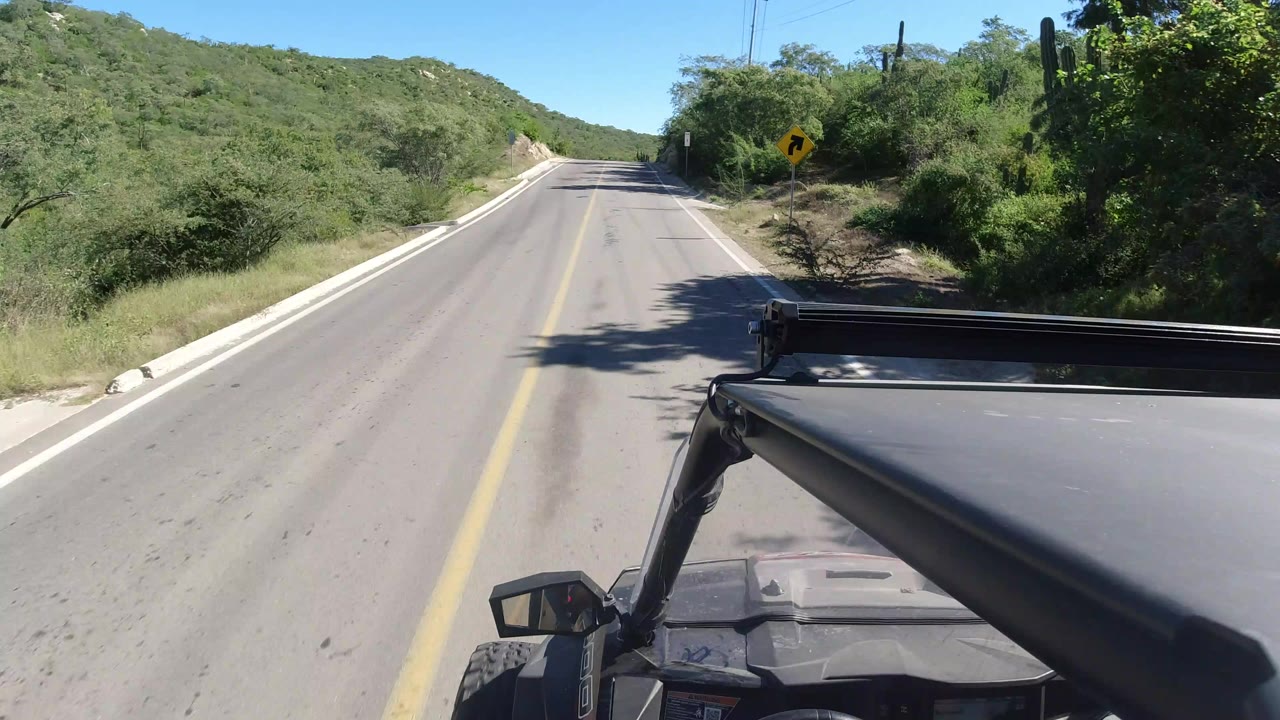 Forward view from a vehicle on a two-lane road in Los Cabos, Mexico, with scrub brush, low green hills, blue sky, and no visible people.