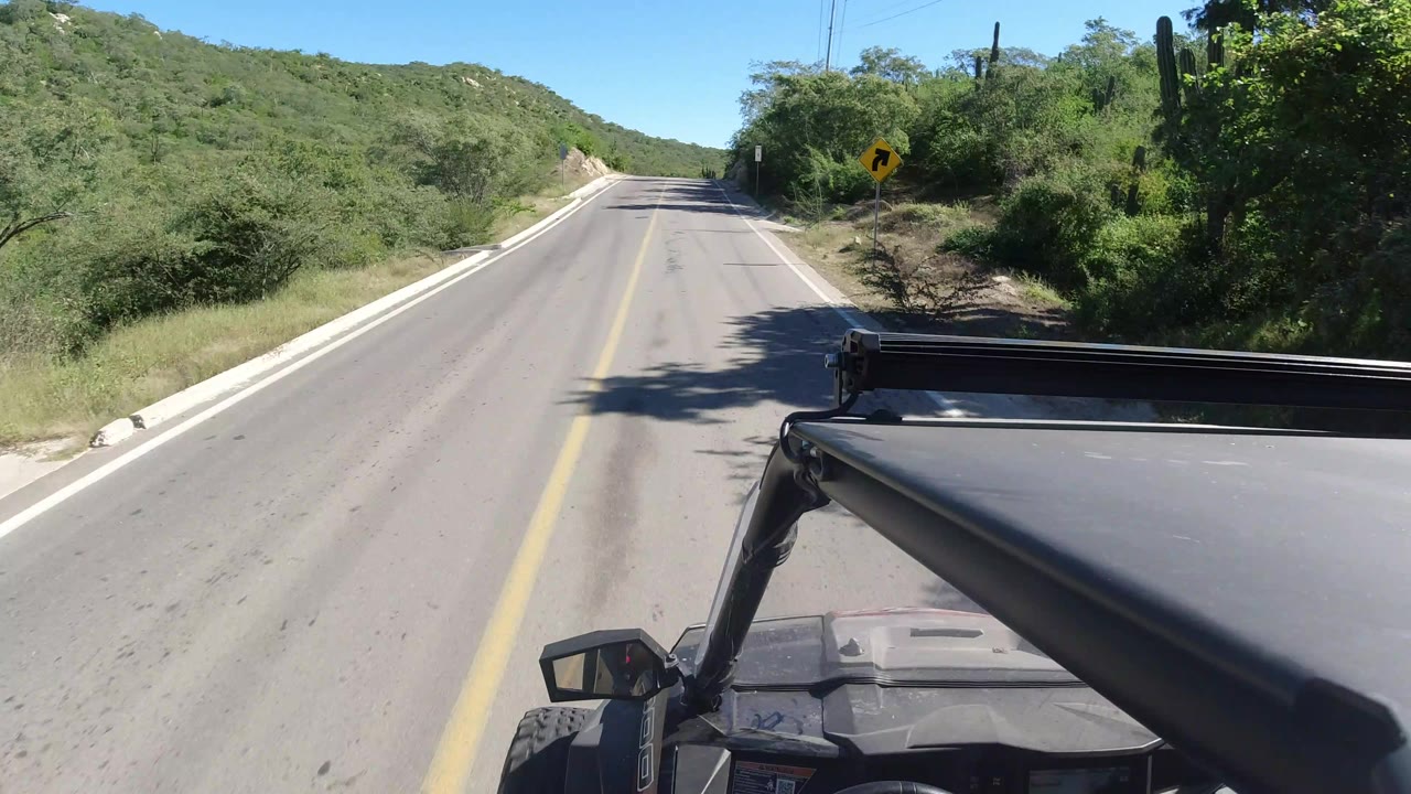The vehicle view continues along a quiet Los Cabos road bordered by scrub vegetation, with the roof edge and roll bar visible in the lower foreground and no visible people.