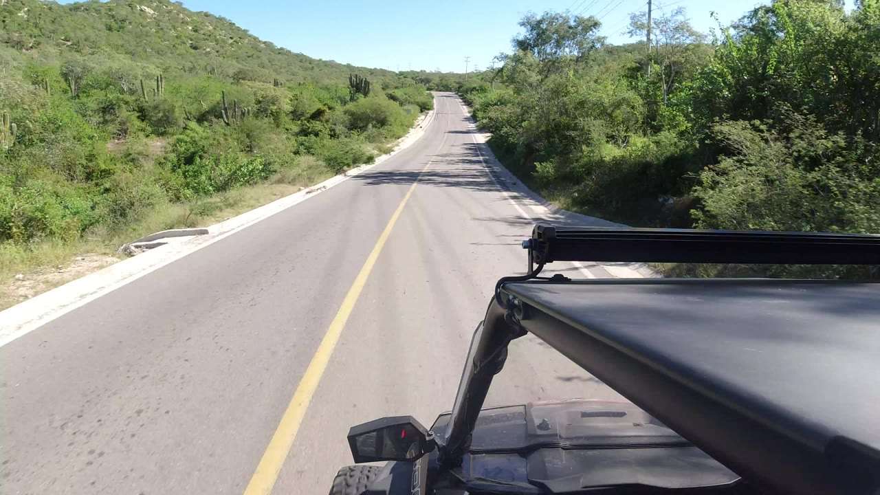 Forward vehicle view on a paved country road in Los Cabos, Mexico, with a yellow center line, green roadside brush, low hills, bright sky, and no visible people.