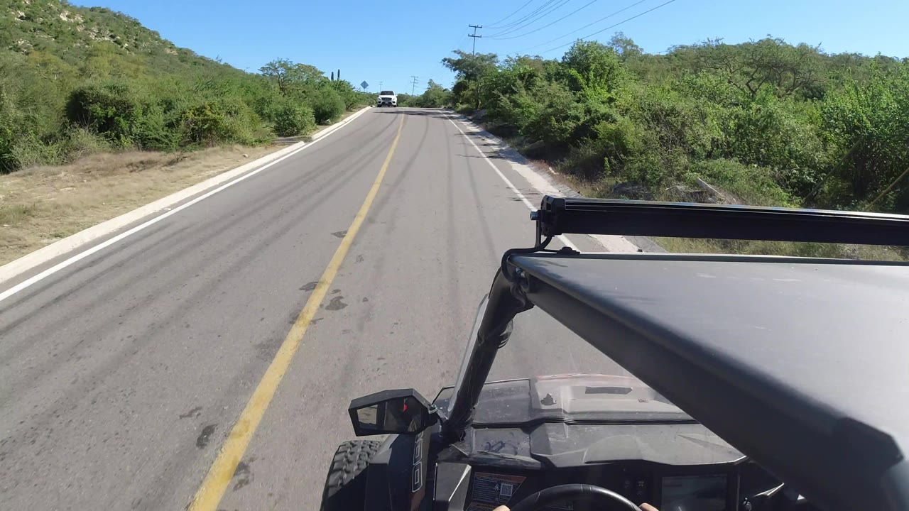 A straight stretch of Los Cabos road runs between dense green vegetation and low hills, with the open vehicle frame visible at lower right and no visible people.