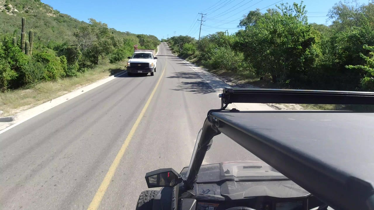 Forward vehicle view on a Los Cabos, Mexico road with an oncoming white truck, green trees on both sides, a visible vehicle roof edge in the foreground, and no visible people.