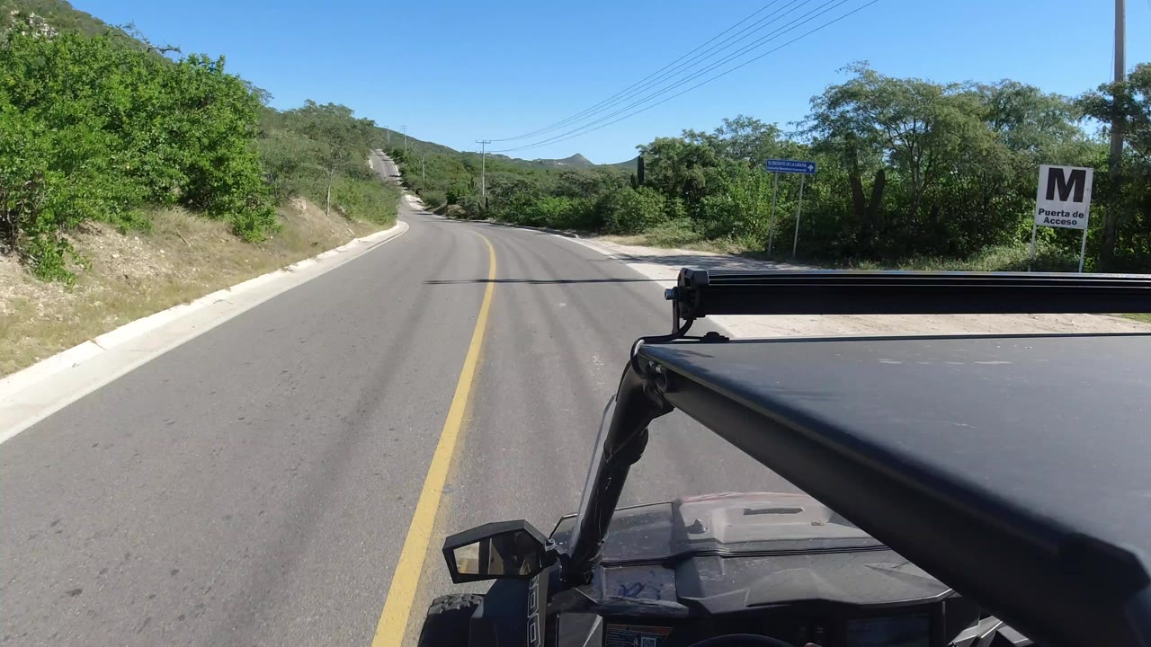 Forward view from a vehicle on a sunlit Los Cabos road with green brush, low hills, a roadside sign, and no visible people.