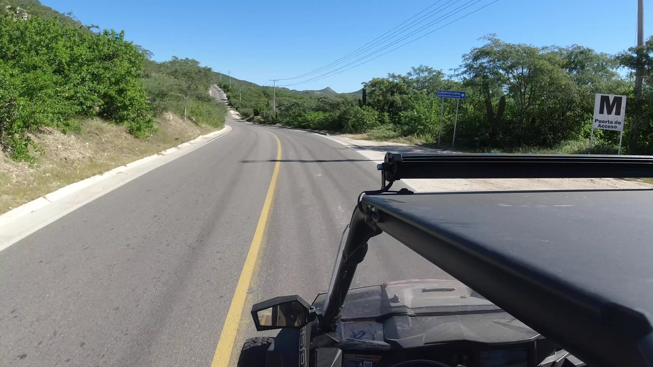 Los Cabos road view from an open vehicle as the yellow centerline leads toward low hills, with green shrubs and no visible people.
