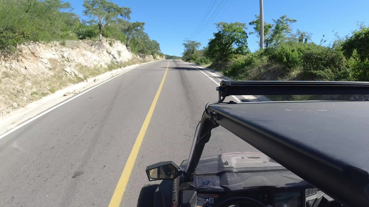 Forward vehicle POV on a two-lane road in Los Cabos, Mexico, with a yellow centerline, pale roadside cut, green brush, blue sky, and no visible people.