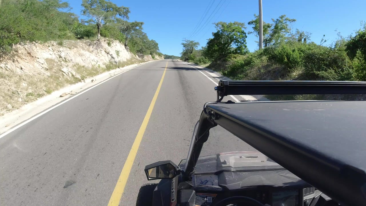 Vehicle-mounted view along the Los Cabos road, with the hood and mirror in the foreground, scrub vegetation beside the pavement, a utility pole, and no visible people.