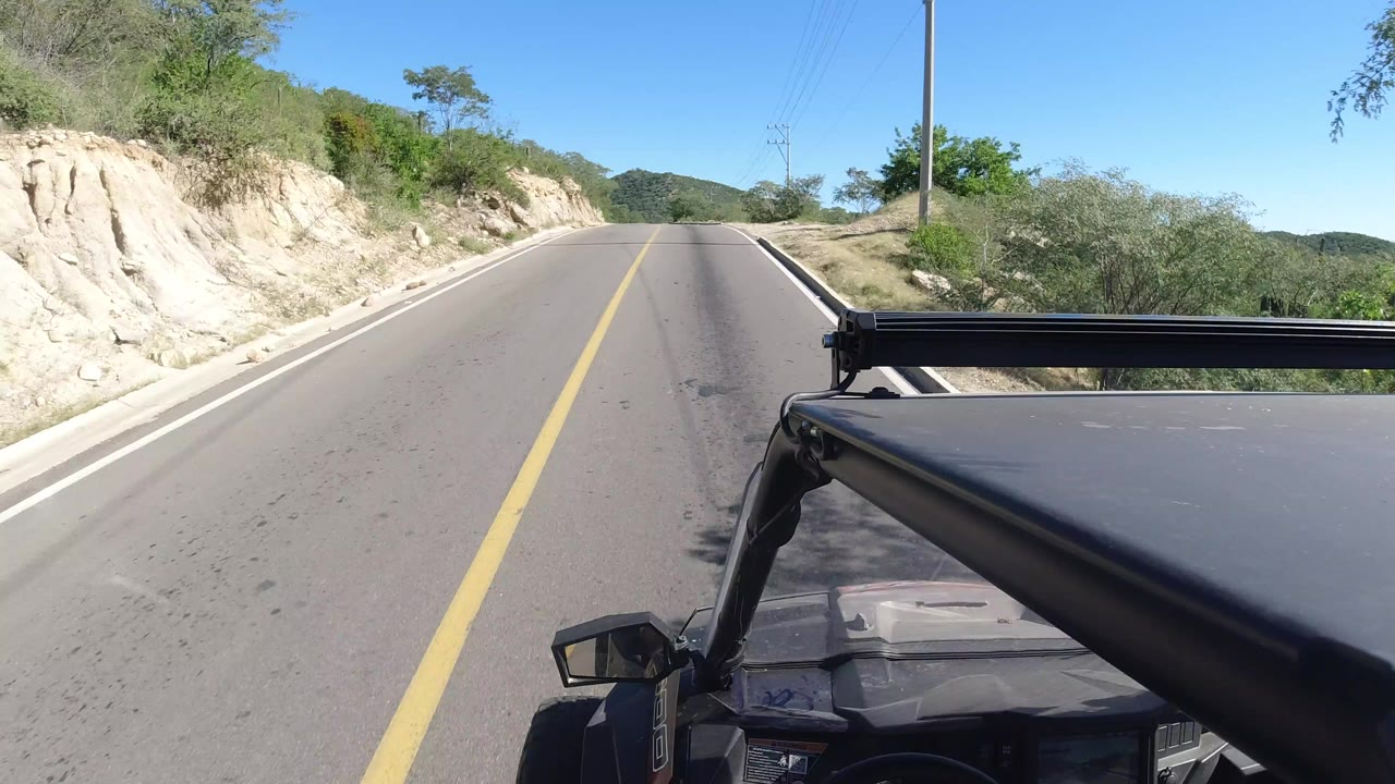 Forward view along an empty road in Los Cabos, Mexico, with a yellow centerline, pale roadside bank, green shrubs, distant hills, blue sky, and no visible people.