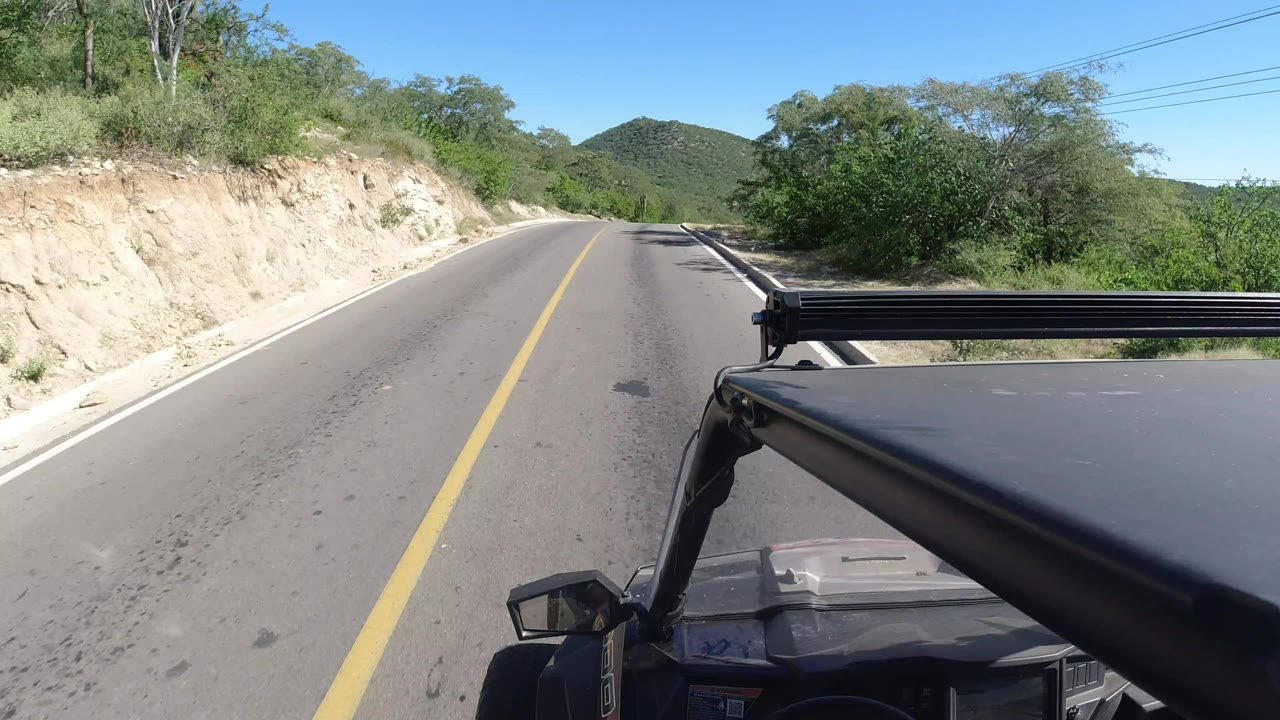 Opening Los Cabos, Mexico vehicle POV with a paved two-lane road, yellow center line, dark car hood and roof rack at lower right, dry cut bank, green brush, low hills, and no visible people.