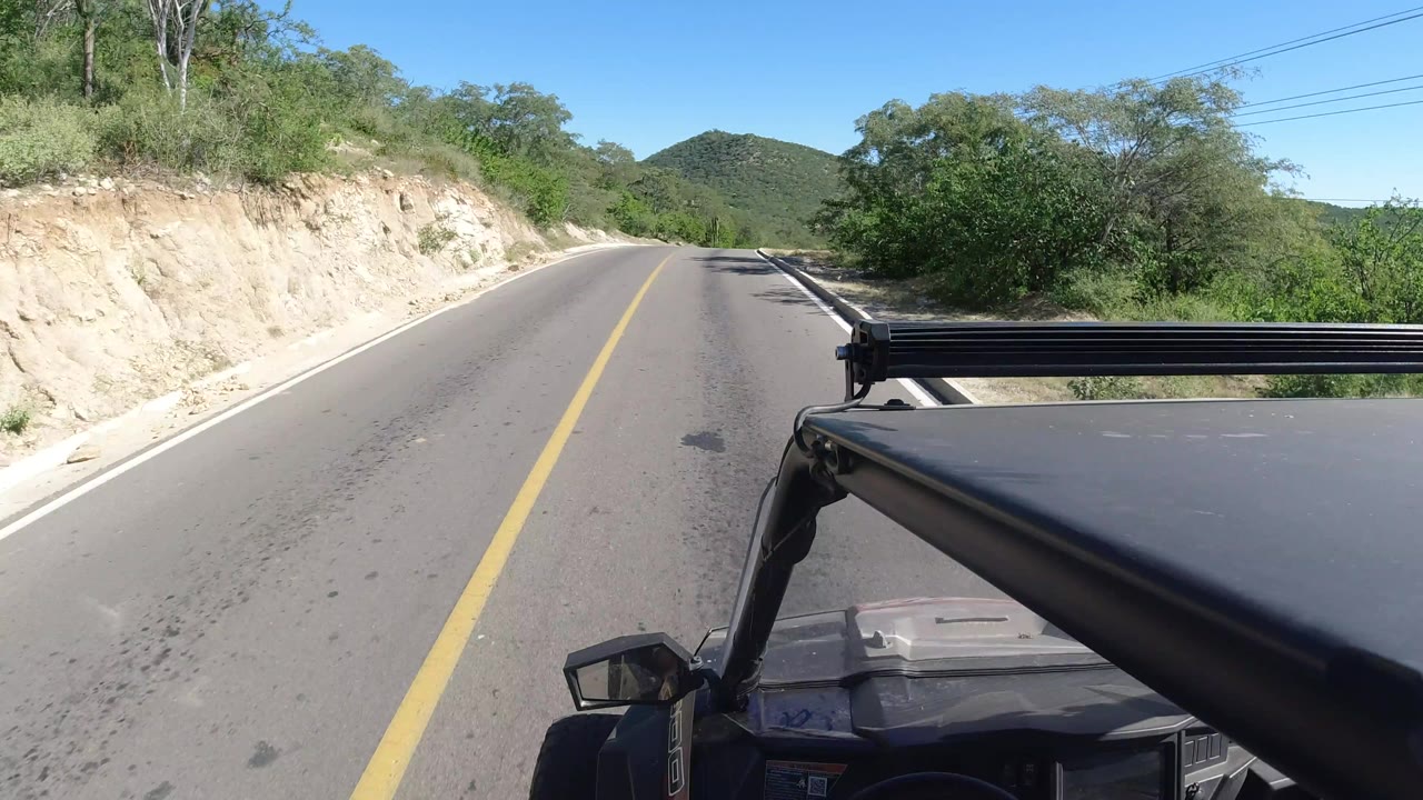Los Cabos, Mexico road view one second in, with the yellow center line leading toward low green hills, roadside scrub on both sides, vehicle hood and rack in the foreground, and no visible people.
