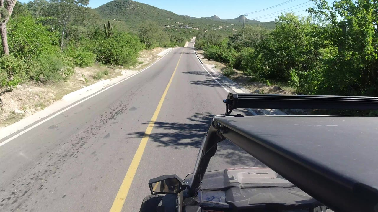 A slightly later road view in Los Cabos, Mexico, keeps the yellow center line centered, with dense green brush on the shoulders, low hills in the distance, vehicle hardware at lower right, and no visible people.
