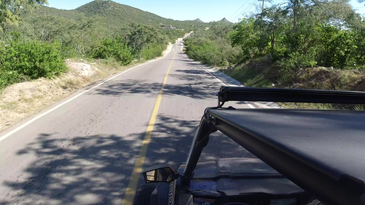 The road continues straight in Los Cabos, Mexico, with the vehicle roof at lower right, dense green brush on both sides, hill slopes ahead, and no visible people.