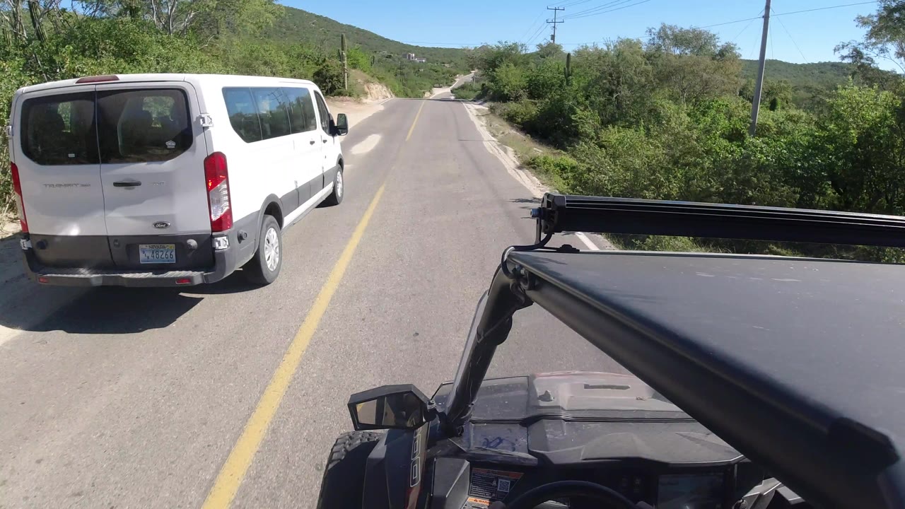The white van remains ahead on the Los Cabos highway as the road curves through green brush and low hills, with the filming vehicle visible at the bottom and no visible people.