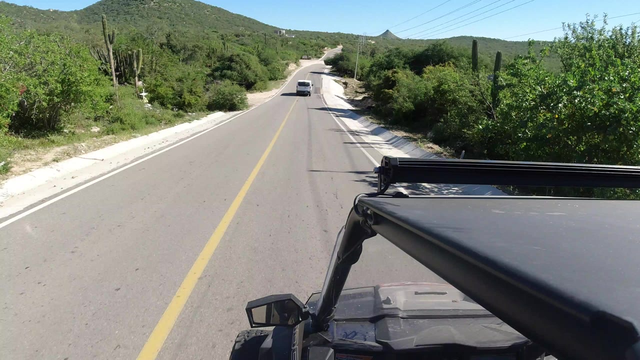 Opening Los Cabos road view with a car ahead on a paved two-lane road, green desert brush, tall cacti, low hills, and no visible people.