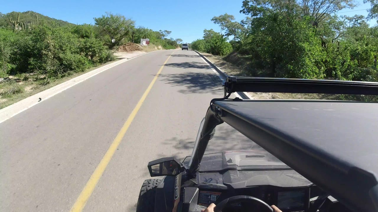 Vehicle POV on a sunny two-lane road with green roadside brush.