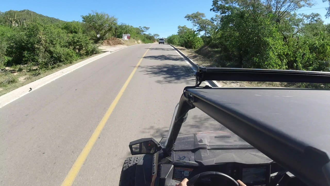 A near-identical forward view continues down the Los Cabos road, with the black vehicle frame at lower right, dense green vegetation, roadside shadows, a distant vehicle, and no visible people.