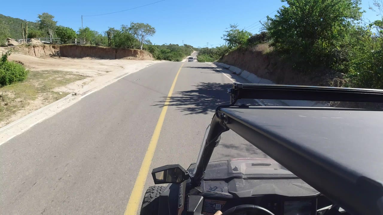 Start of the Los Cabos, Mexico road clip, showing a paved two-lane road, sandy shoulders, green roadside trees, and part of the vehicle in the lower right with no visible people.