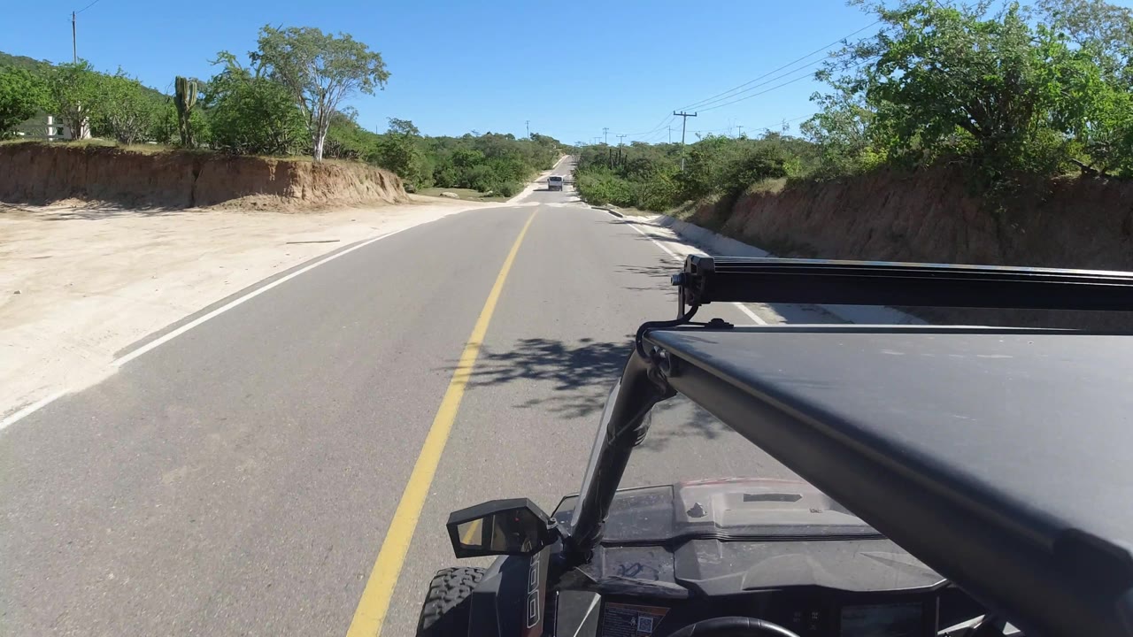 The Los Cabos, Mexico roadway continues straight ahead with a pale sandy pullout on the left, green trees on both sides, overhead utility lines, and no visible people.