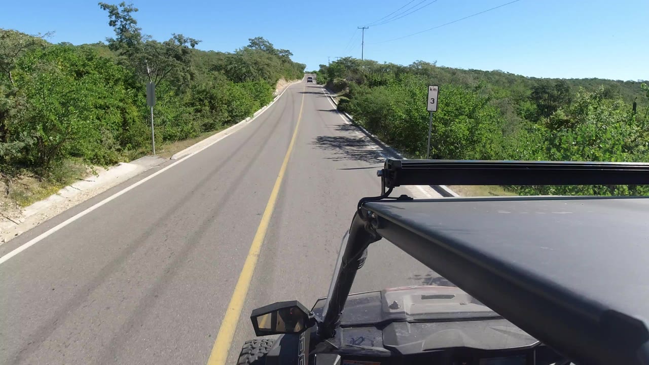 Forward view down a two-lane road bordered by green brush from an open vehicle.