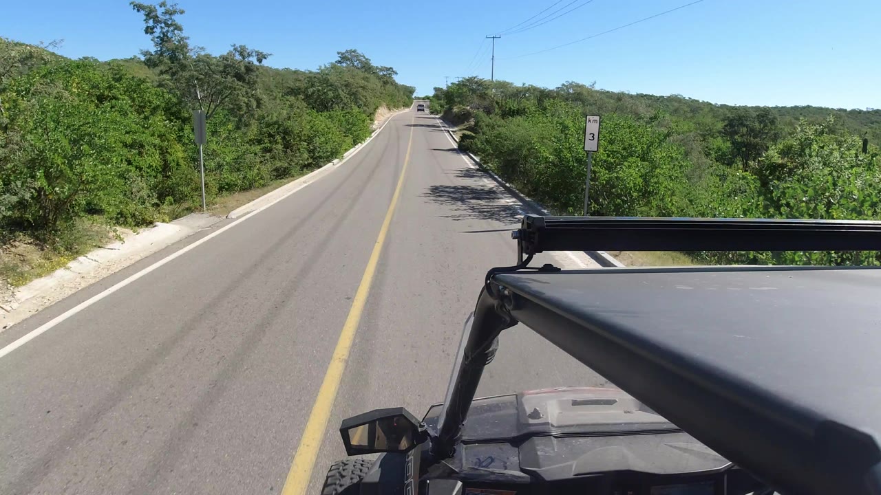 Early in the Los Cabos, Mexico drive, the centered road continues past thick green vegetation with the vehicle roof and roll bar filling the lower right foreground and no visible people.