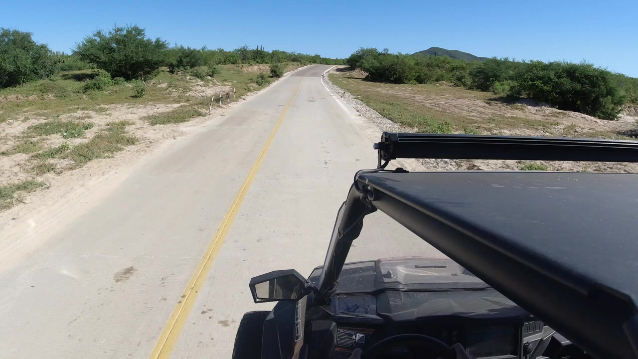 A slightly farther moment along the Los Cabos road shows the yellow line leading toward a bend, with the dark vehicle roof and roof bar cutting across the lower right and no visible people.