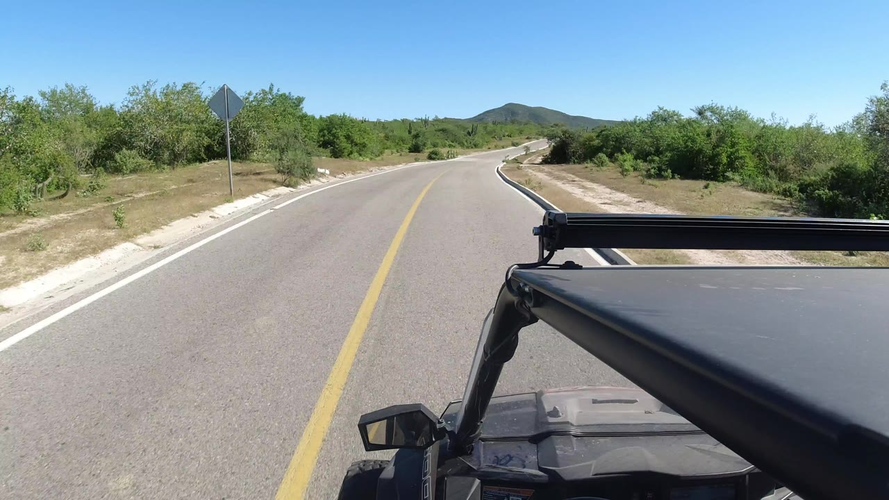 Start of the Los Cabos, Mexico road clip with a two-lane paved road curving ahead, green shrubs on both sides, low hills in the distance, and no visible people.