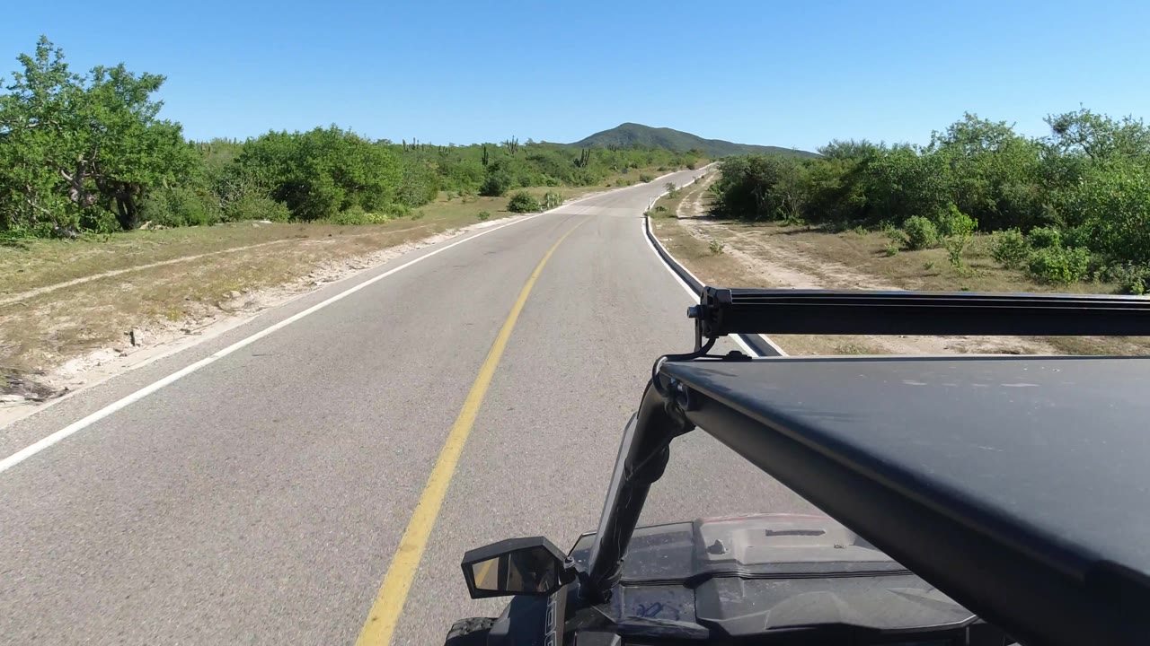Open paved road with green brush, distant hills, and vehicle hood at right