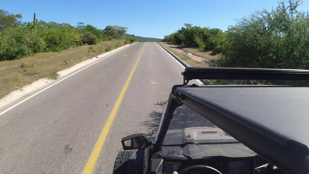 Opening frame of an empty Los Cabos road from a vehicle-mounted viewpoint, with a yellow center line, green brush, distant hills, blue sky, and no visible people.