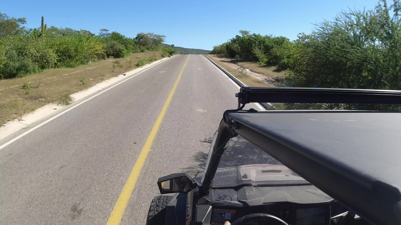 Second frame of the Los Cabos driving POV, showing the vehicle progressing along a straight clean road with dense green brush, low hills, and no visible people.