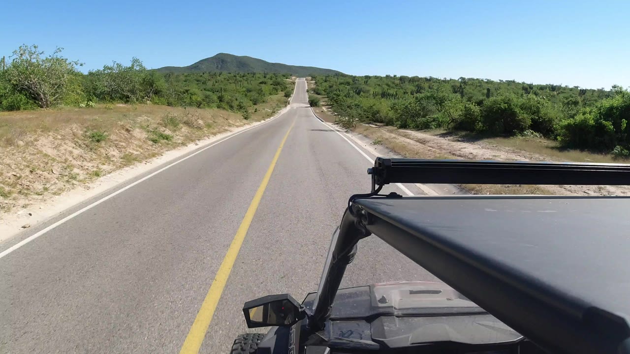 Forward view down an empty paved road in Los Cabos, Mexico, with a black off-road vehicle roof in the foreground, green shrubs on both sides, low hills ahead, and no visible people.