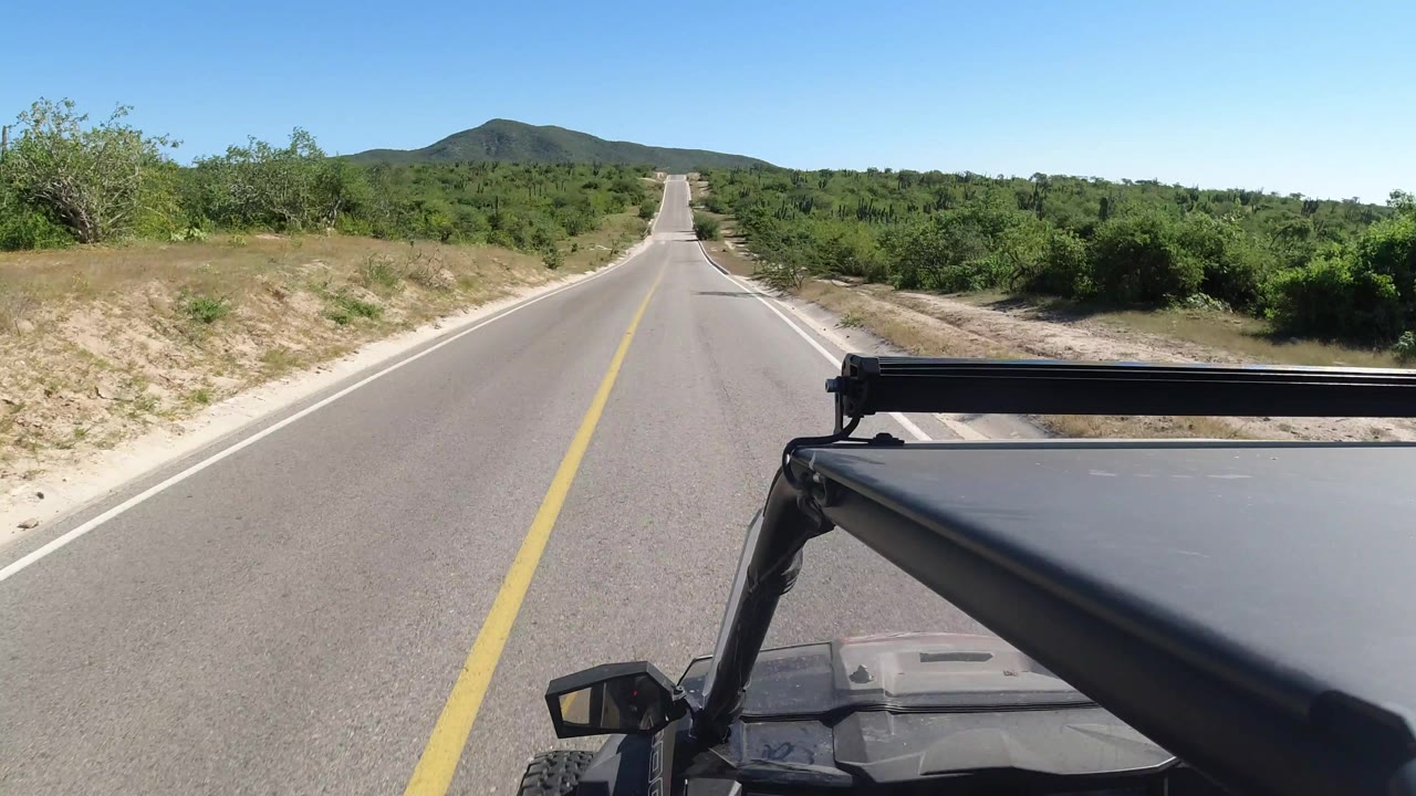 The road continues straight through Los Cabos scrubland, with the vehicle roof and roll bar low in frame, sandy shoulders, distant hills, and no visible people.