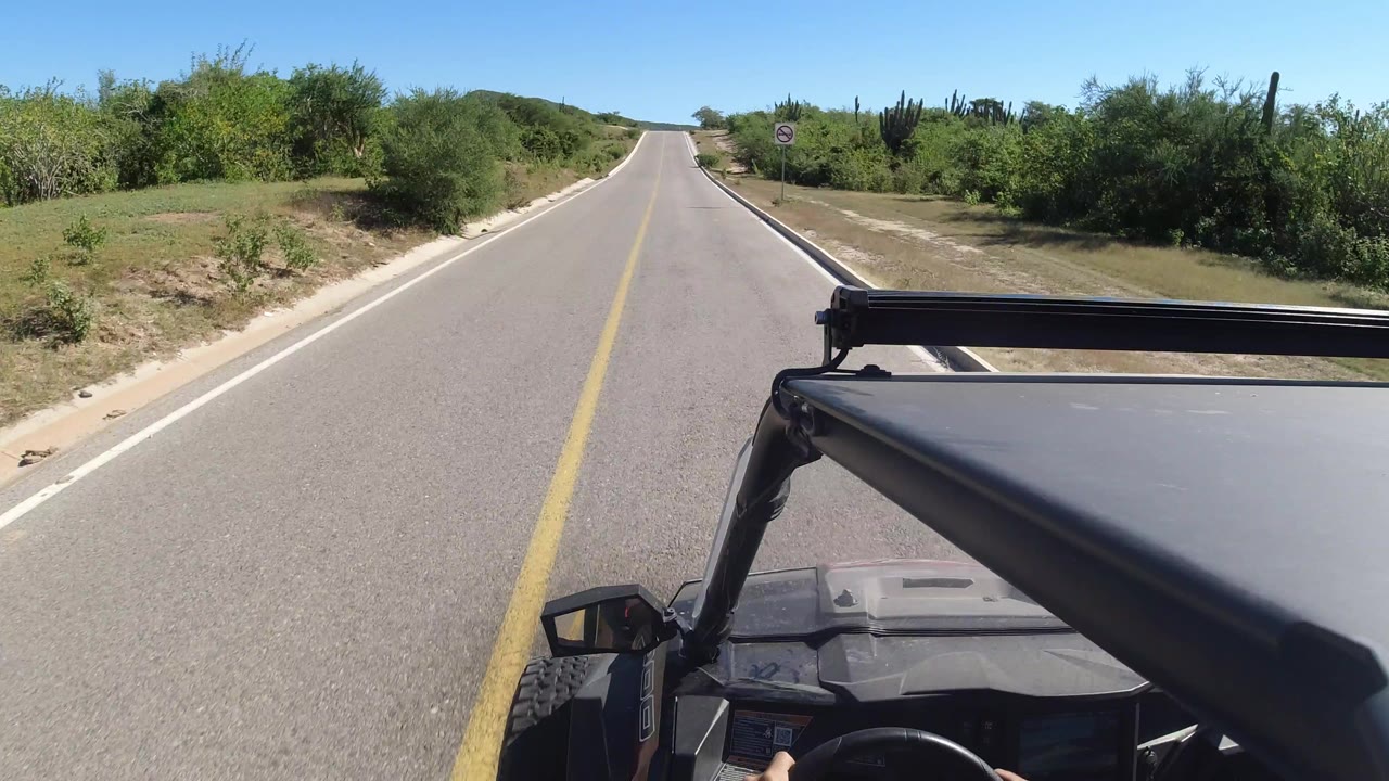 The road continues through low green vegetation in Los Cabos, with the vehicle's dark hood and roof rack along the lower right and no visible people.