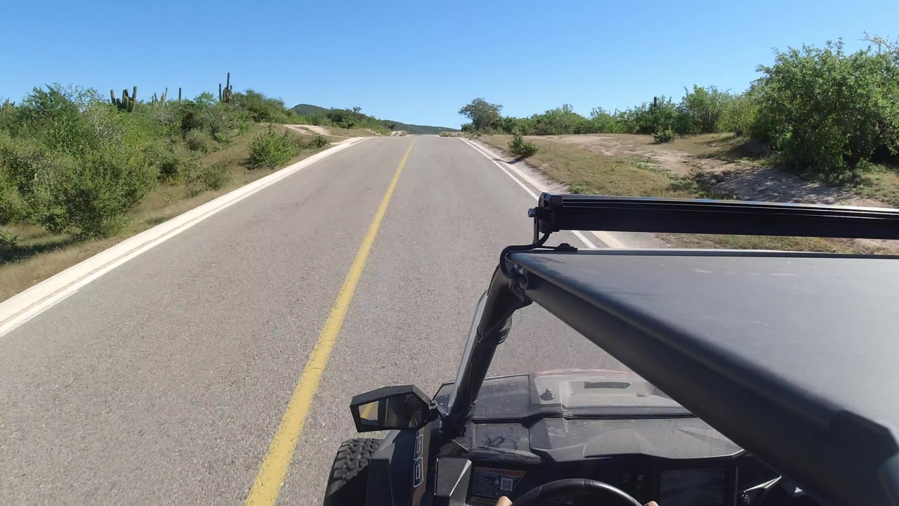 Start of a Los Cabos road drive seen from a black vehicle, with a yellow center line, green shrubs, cactus, blue sky, and no visible people.