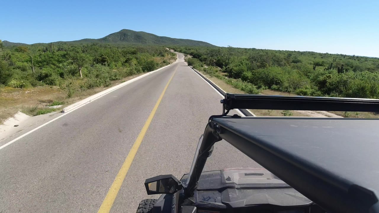 Forward-facing vehicle view on a Los Cabos road, with the lane centered, low green plants on both sides, mountains ahead, and no visible people.