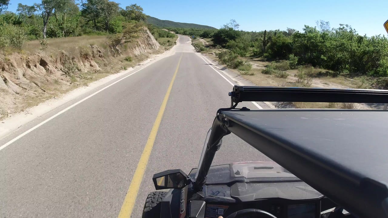 Paved road seen from an open vehicle with shrubs and low hills ahead.