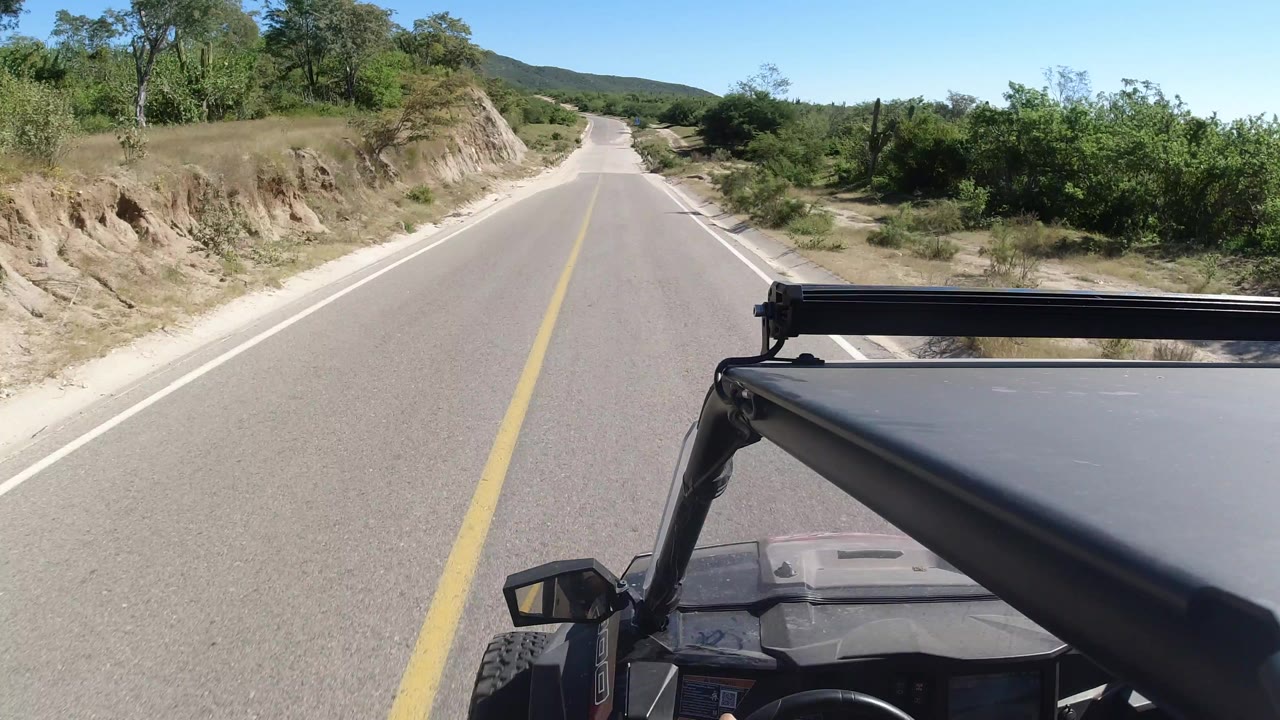 Vehicle POV along a straight Los Cabos road, showing the hood and mirror at the bottom, a pale road shoulder, dense green scrub, distant hills, and no visible people.