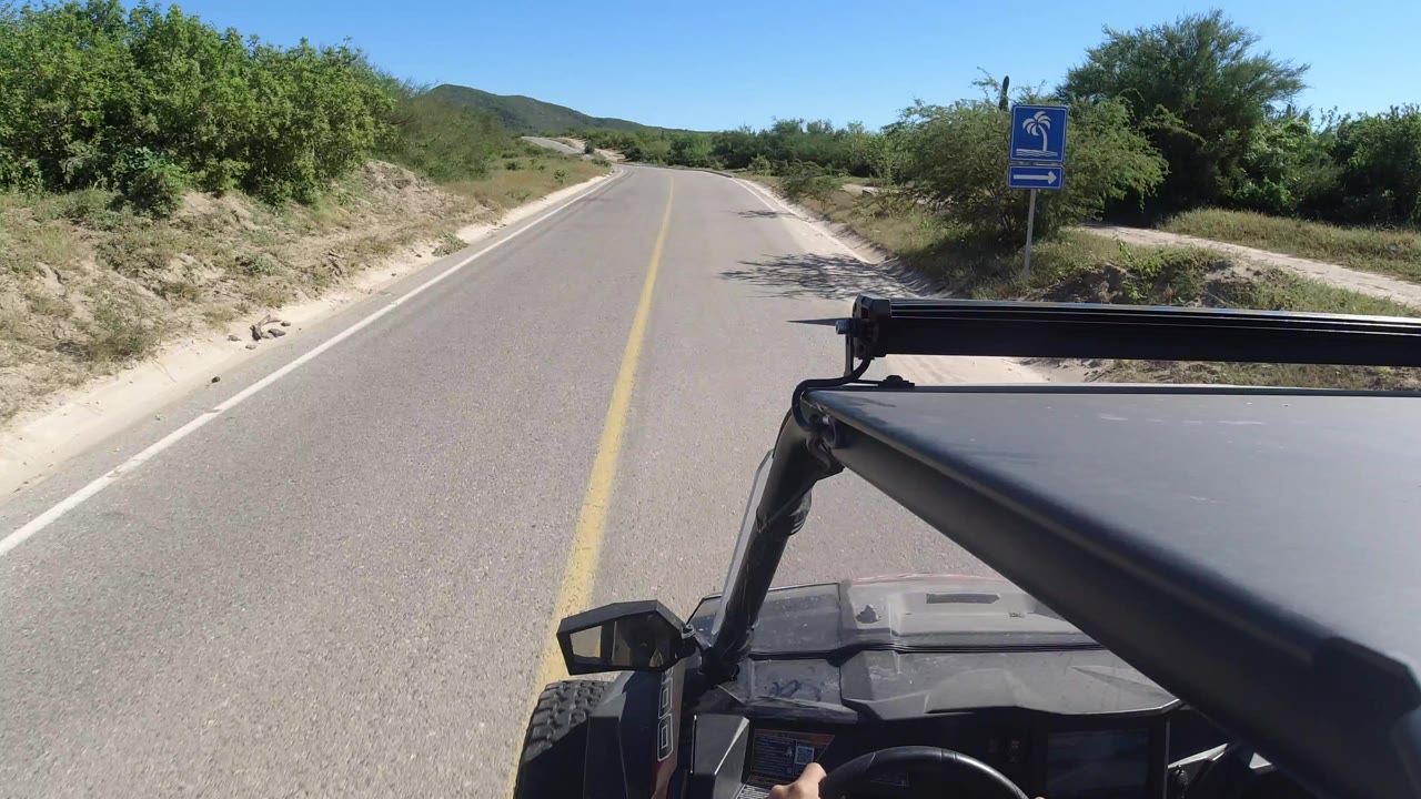 Start of a Los Cabos road drive from an open vehicle, with a yellow center line, green brush, a roadside sign, vehicle hardware in the foreground, and no visible faces.