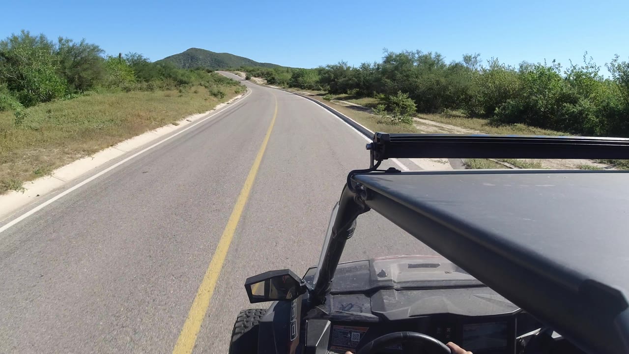 Empty road curving through green roadside vegetation with vehicle parts at right.