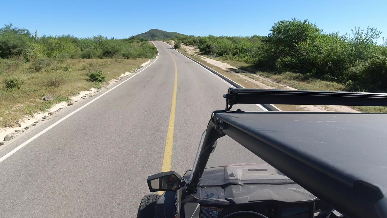 Opening view in Los Cabos, Mexico, from beside a black SUV roof bar looking down an empty two-lane road toward low hills, with no visible people.