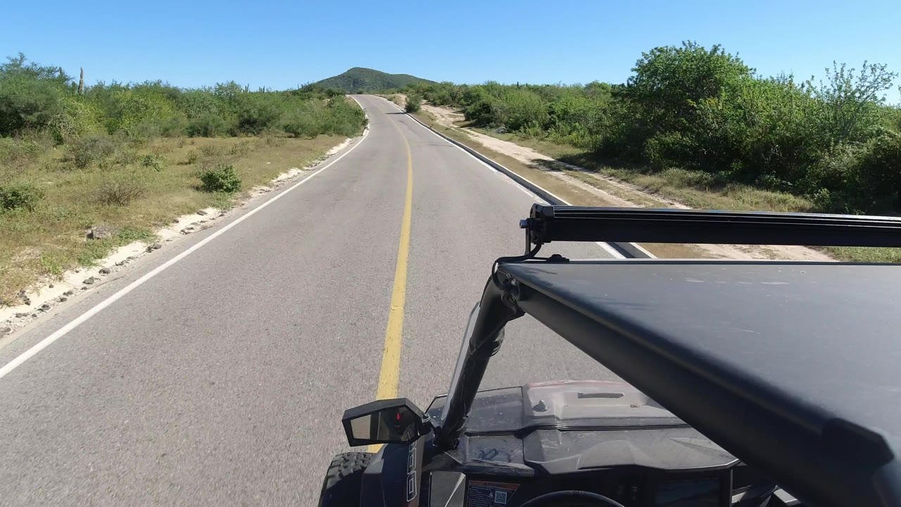 Los Cabos highway view with the SUV roof and rack occupying the lower right foreground, green brush on both sides, and a clear blue sky with no visible people.