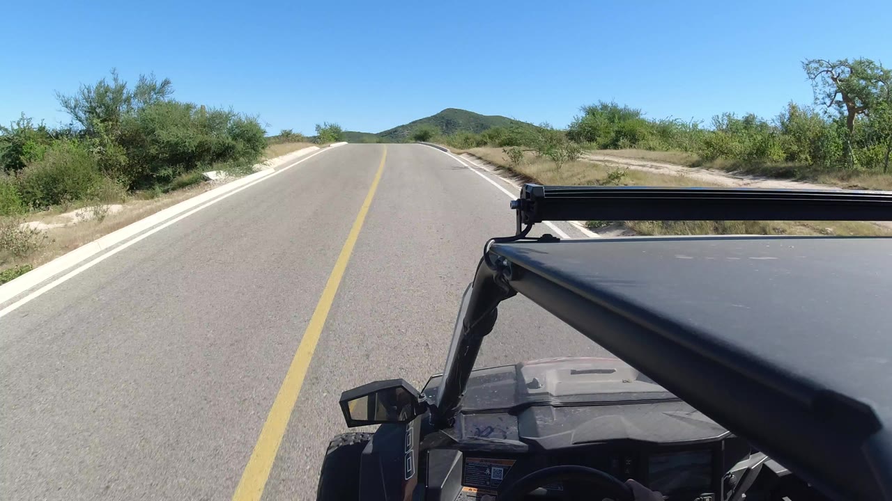 Forward road view in Los Cabos, Mexico, with a yellow center line, low roadside trees, distant hills, bright blue sky, and no visible people.