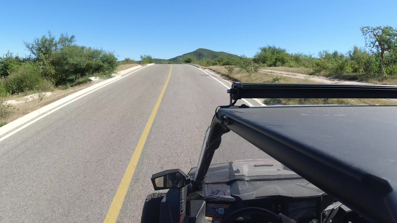 Los Cabos road POV showing the lane stretching toward low hills, green scrub on both sides, a black vehicle roof bar in front, and no visible people.
