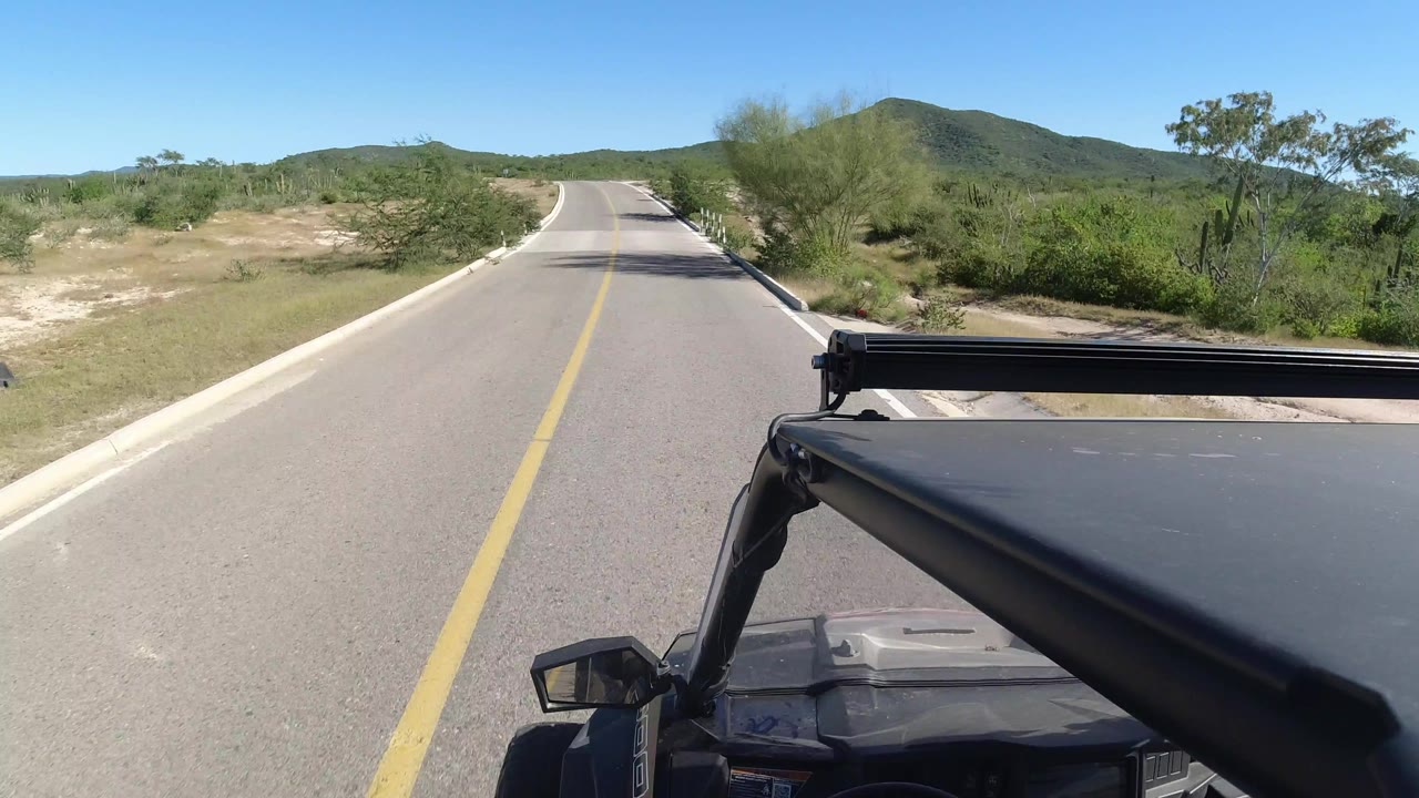 Vehicle view down an empty paved road toward low green hills.